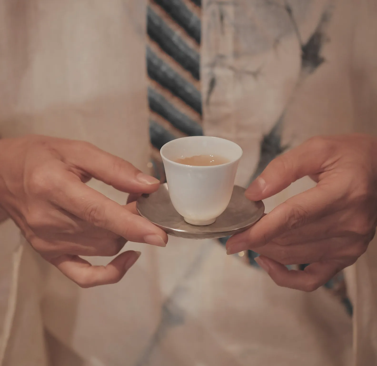 Two hands holding a small white cup on a saucer, against a linen robe