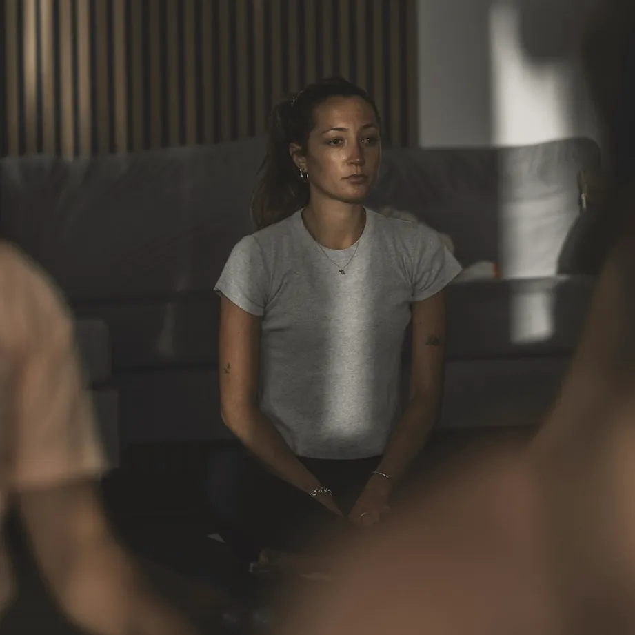 A woman sitting cross legged in a dim room with timber slats, others in the class blurred in front of her