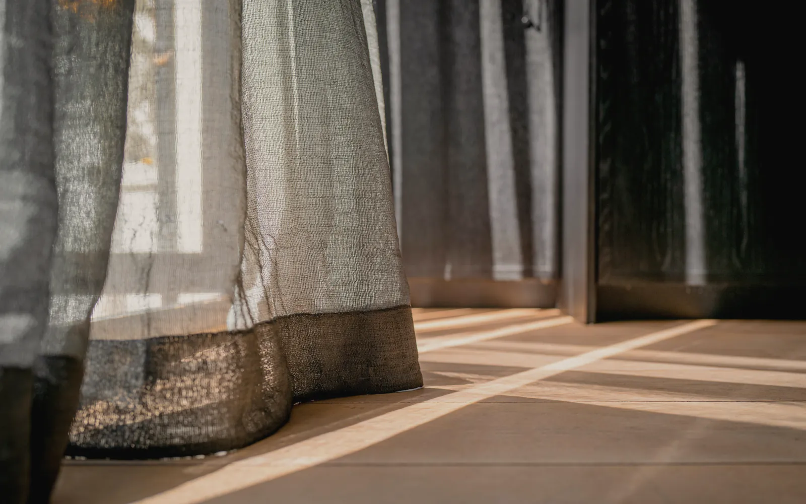 A linen curtain falling to a pale floor, with low sun lying across the boards