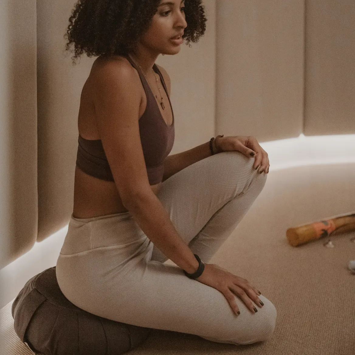 A woman sitting on a floor cushion in a softly lit room, one hand on her knee