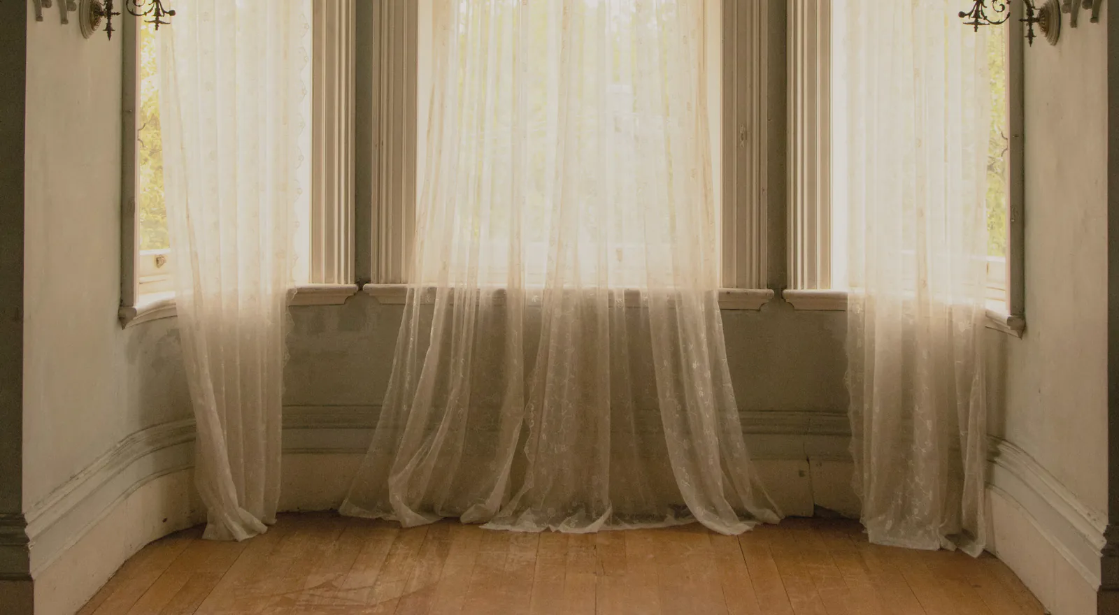 Lace curtains drawn across the tall sashes of a bay window in an old house, above a timber floor