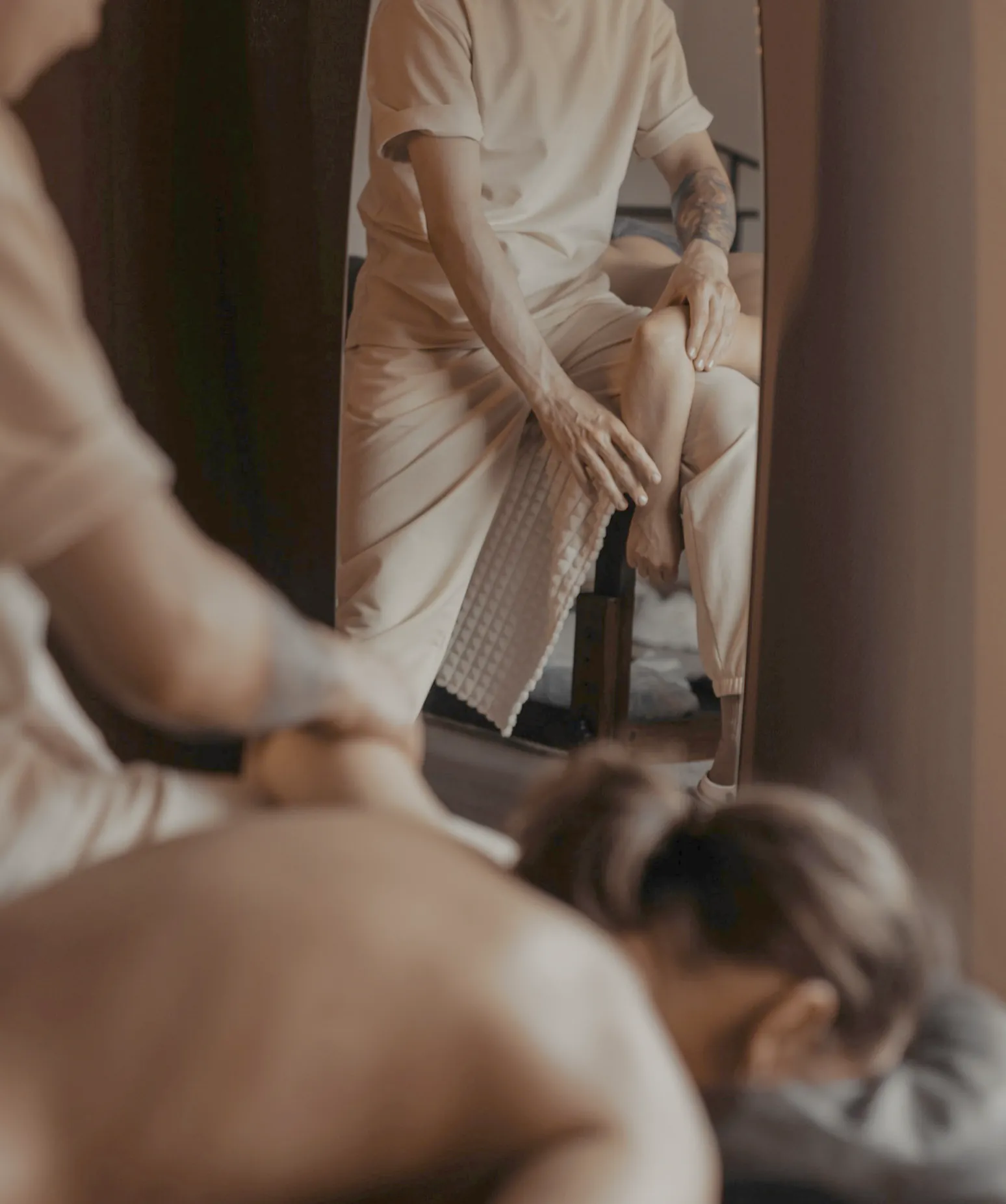 A massage therapist working along the back of a client lying face down, a tall mirror behind them, in warm lamp light
