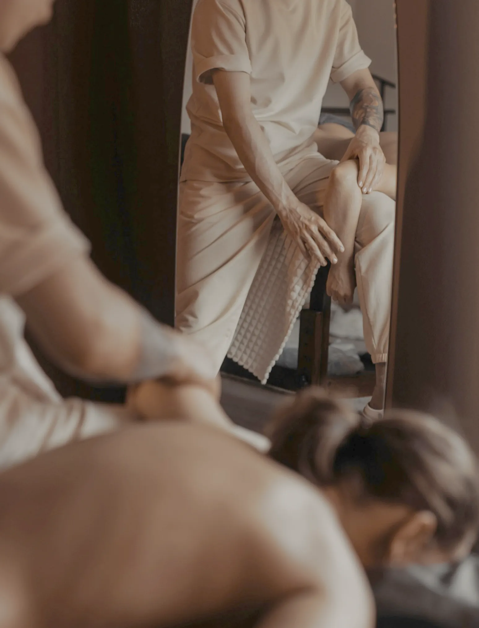 A massage therapist working along the back of a client lying face down, a tall mirror behind them, in warm lamp light