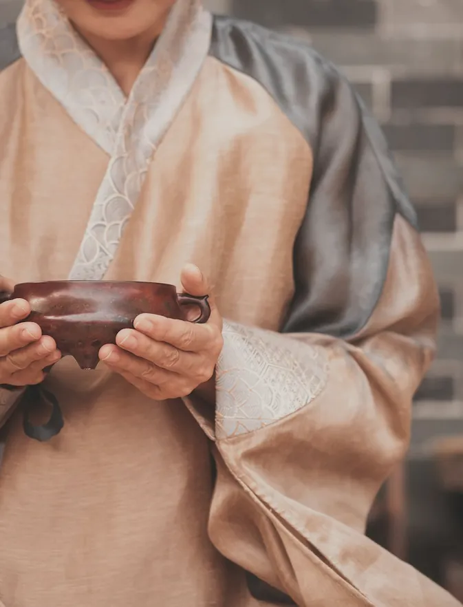Two hands holding a small clay bowl of tea against a linen robe