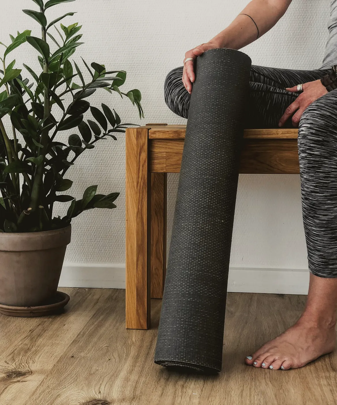 Someone sitting on a timber bench holding a rolled mat, a potted plant beside them