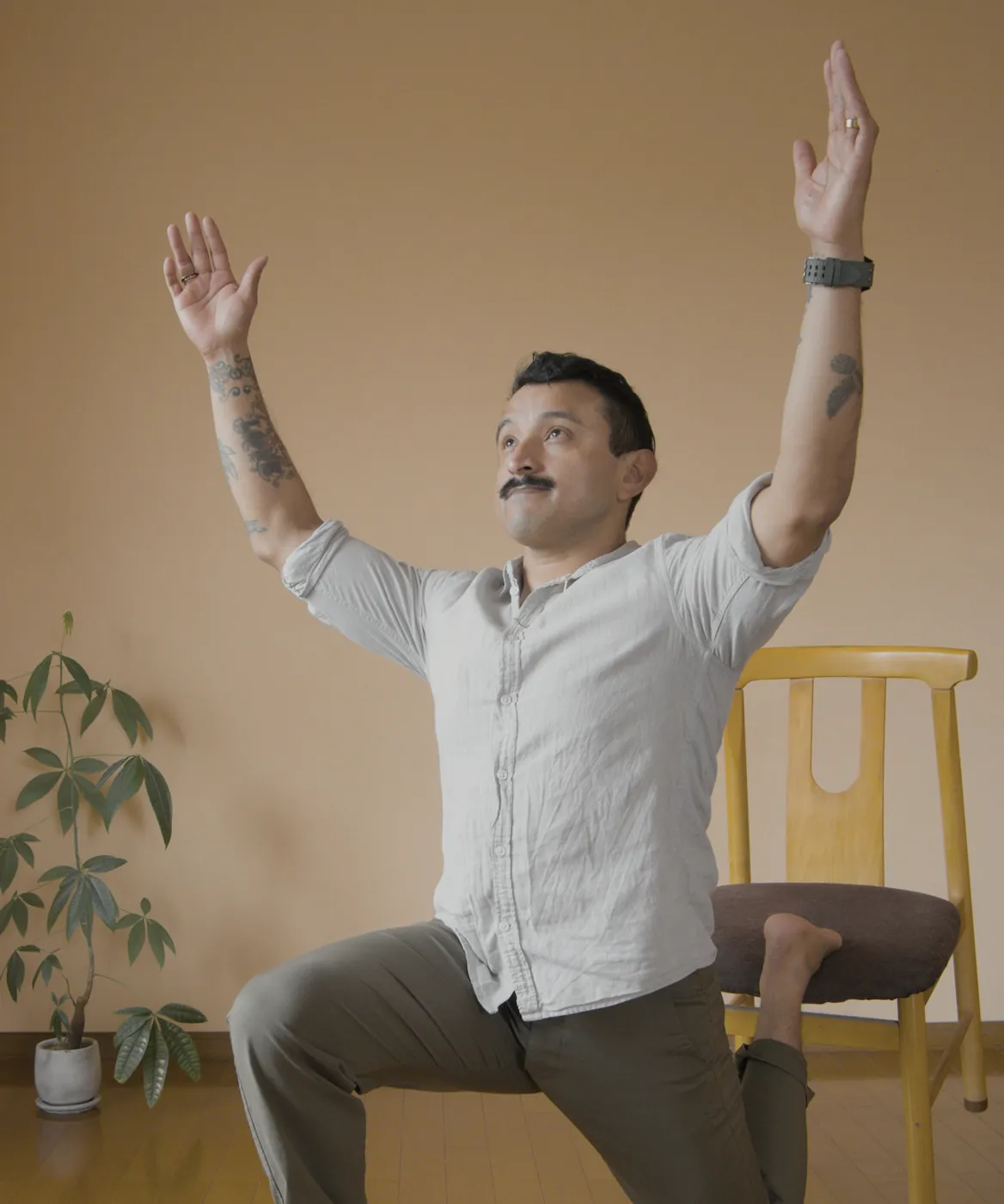 A man in a slow lunge with his back foot resting on a timber chair and both arms raised, in a warm room