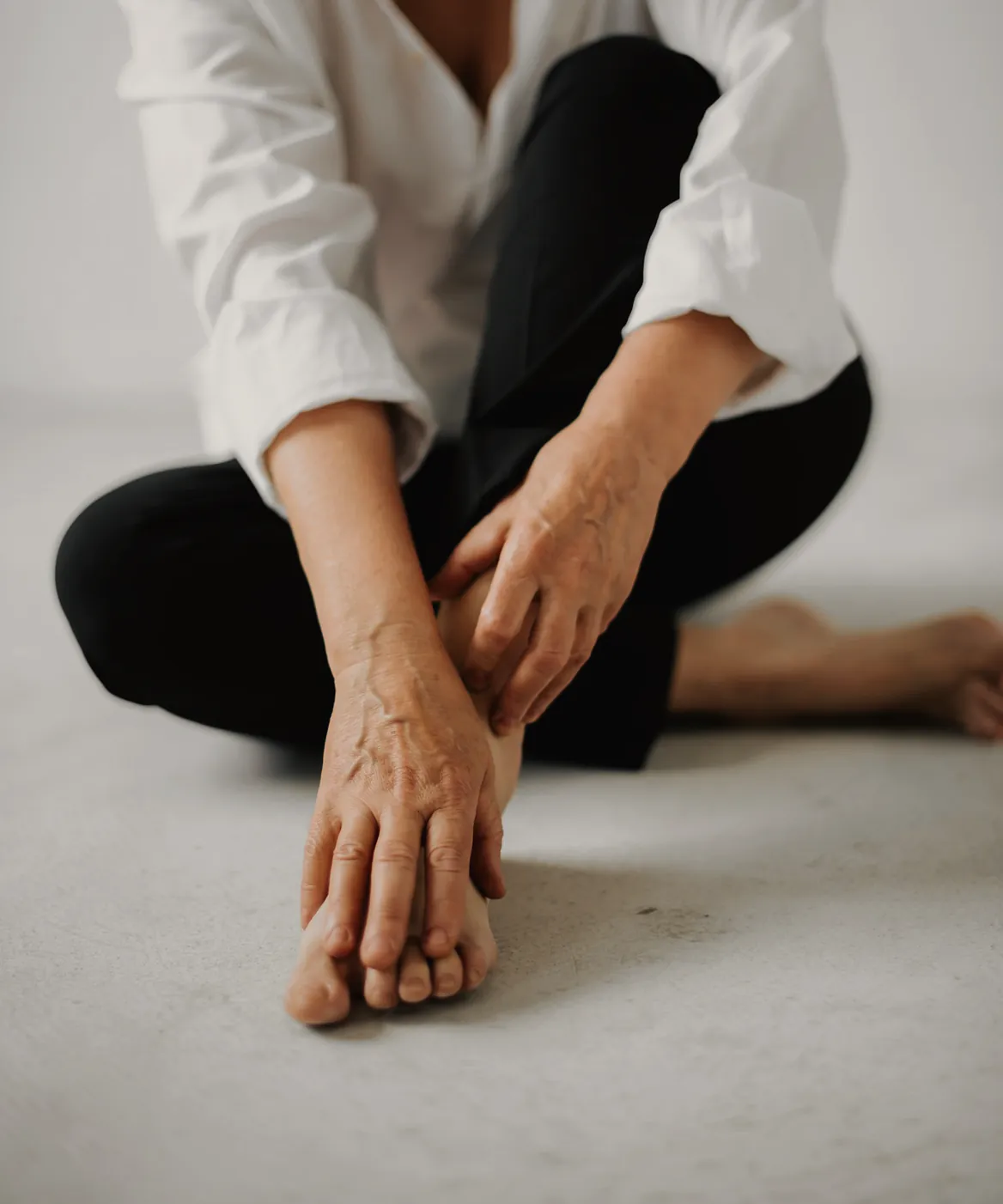 A woman in a white shirt sitting on the floor, holding one foot and pausing