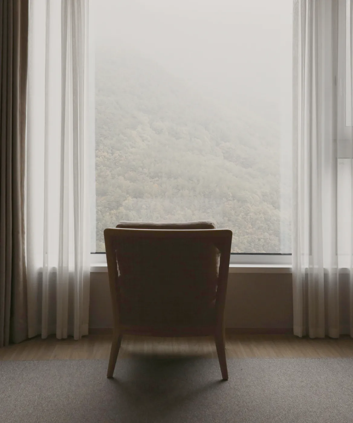 A chair turned to a tall window between long curtains, misty hills outside