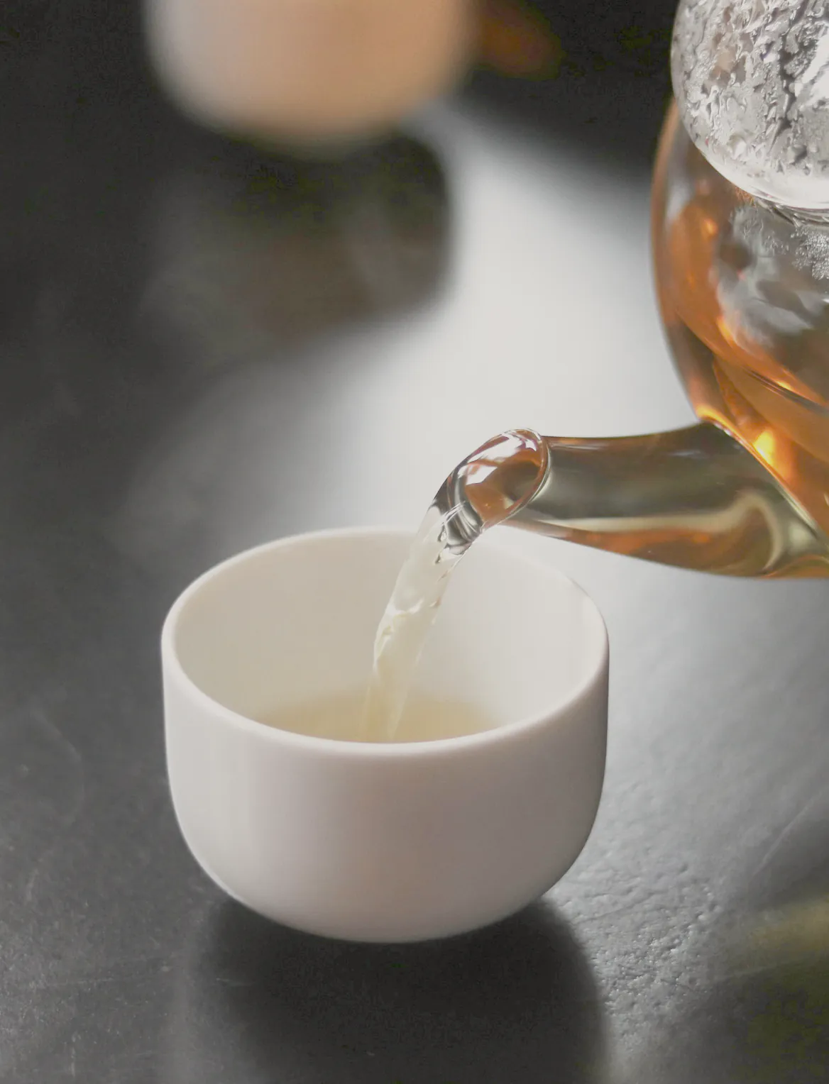 Pale tea poured from a glass teapot into a small white cup on a dark table