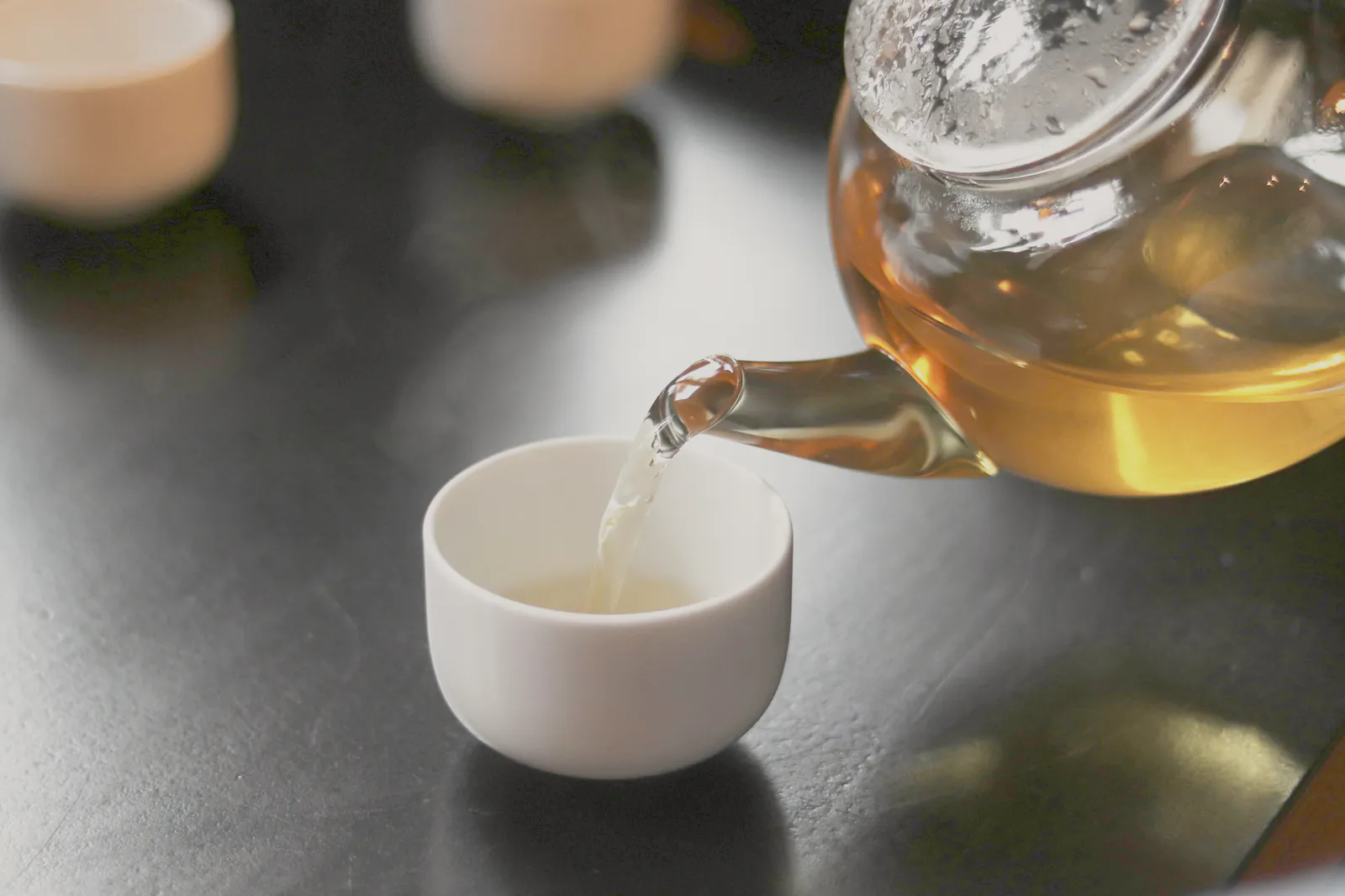 Pale tea poured from a glass teapot into a small white cup on a dark table