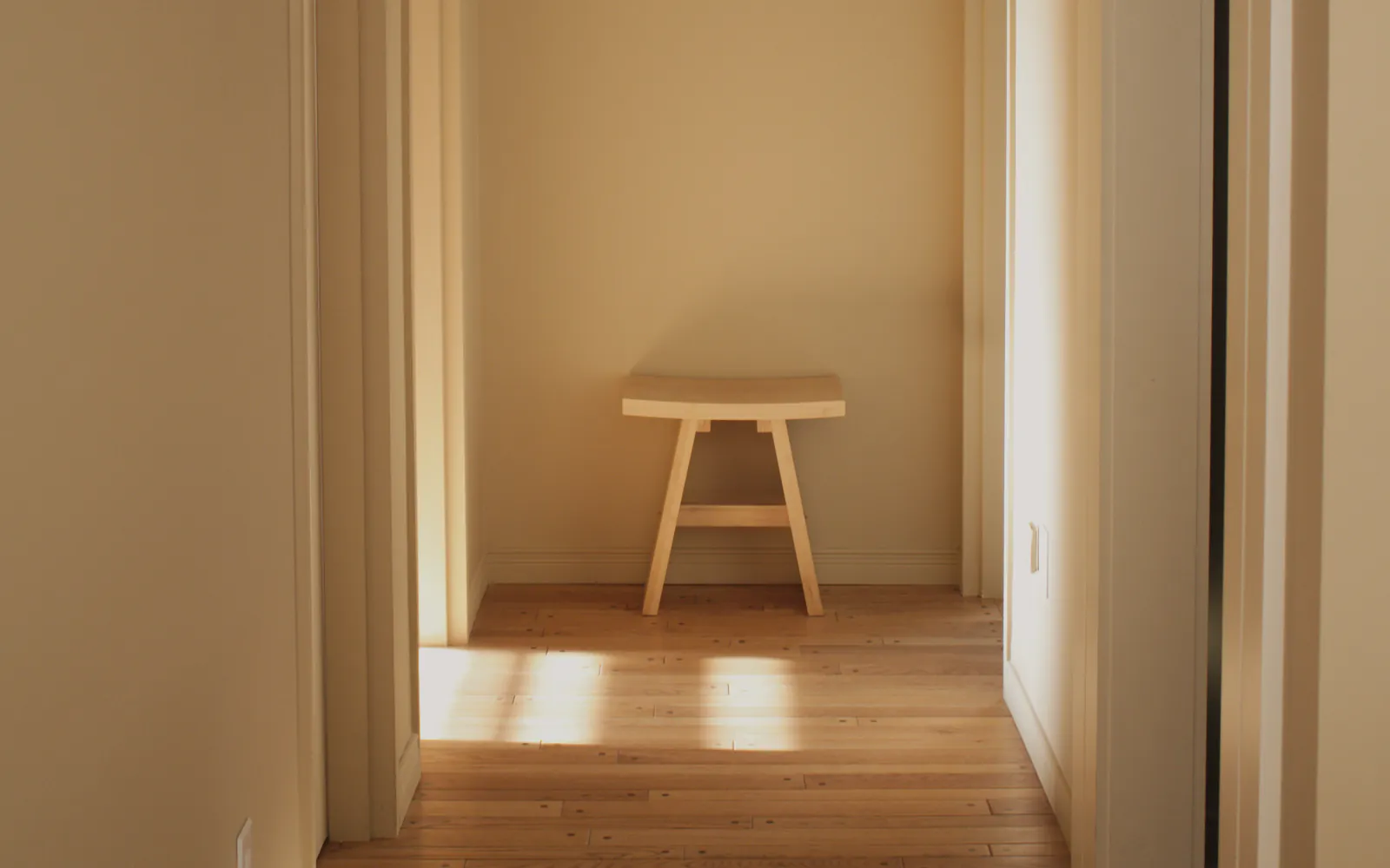 A quiet hallway with timber boards and warm painted walls, sun lying in patches on the floor and a small stool at the end