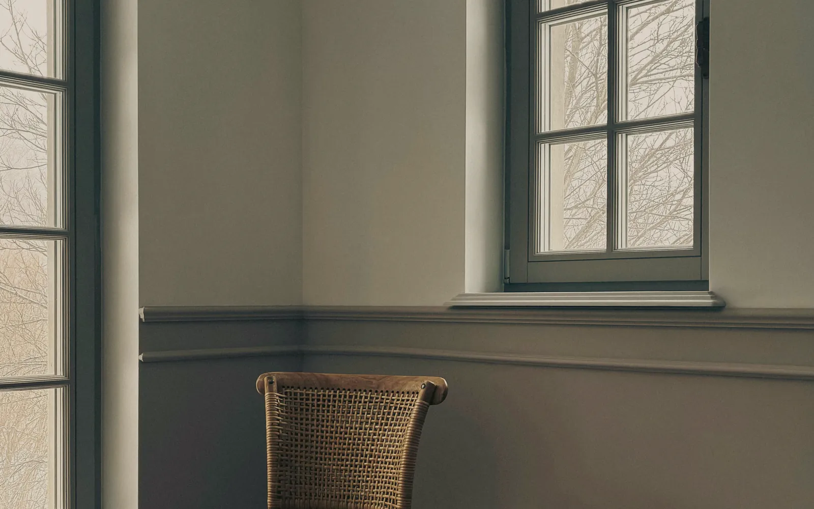 A plain grey green room in soft daylight, two tall windows onto bare trees and a cane chair against the wall
