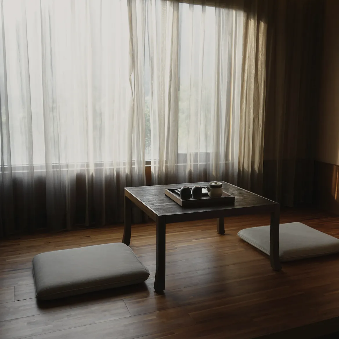 A low timber table set with a tea tray between two floor cushions, sheer curtains drawn across the window