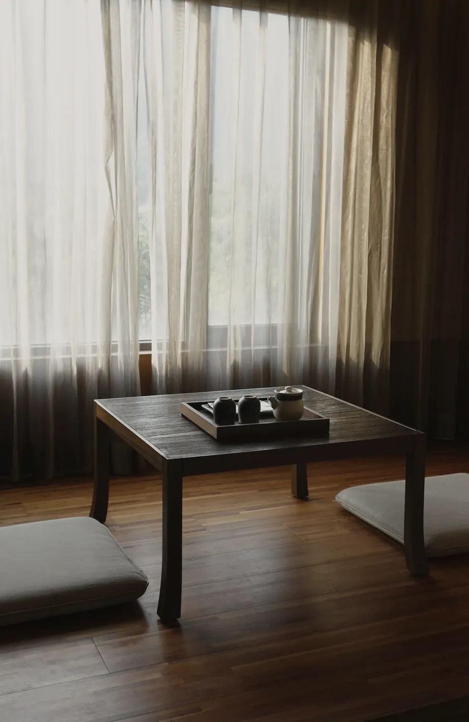 A low timber table set with a tea tray between two floor cushions, sheer curtains drawn across the window
