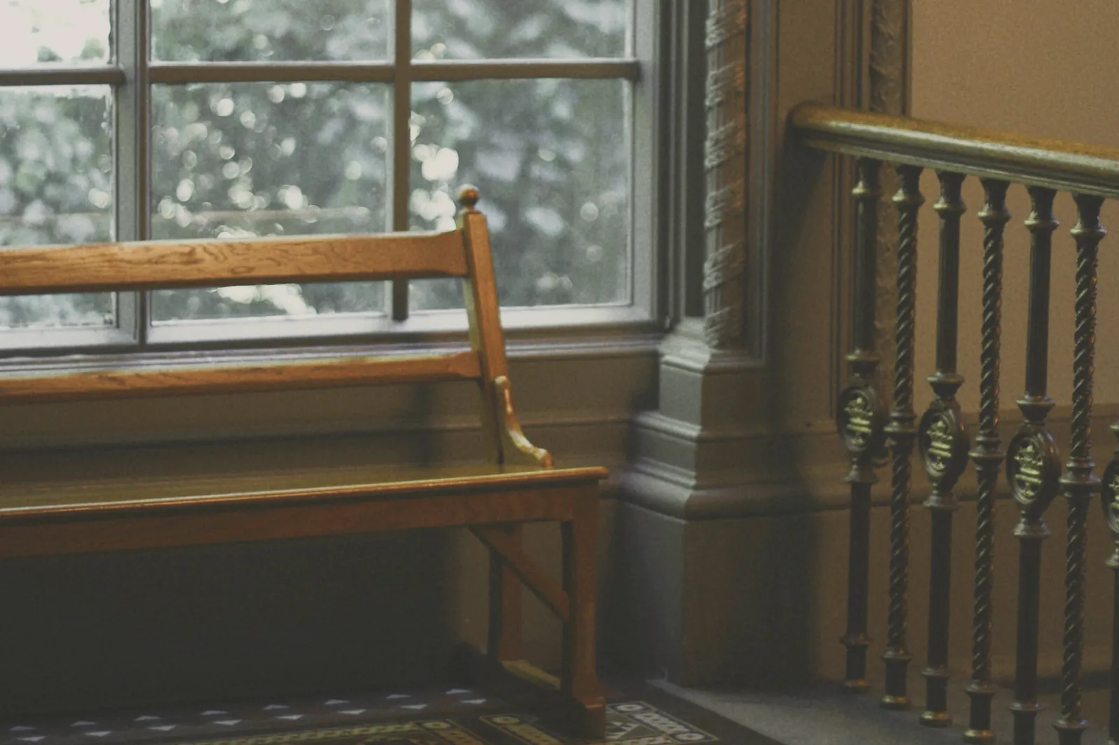 A timber bench under a tall sash window onto green trees, beside an iron balustrade and a tiled floor