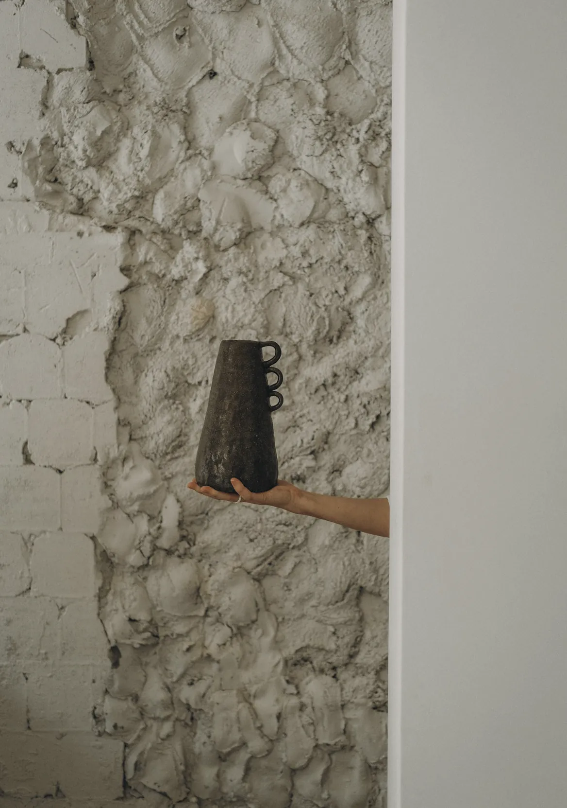 A hand holding a small black clay cup beside a rough plaster wall